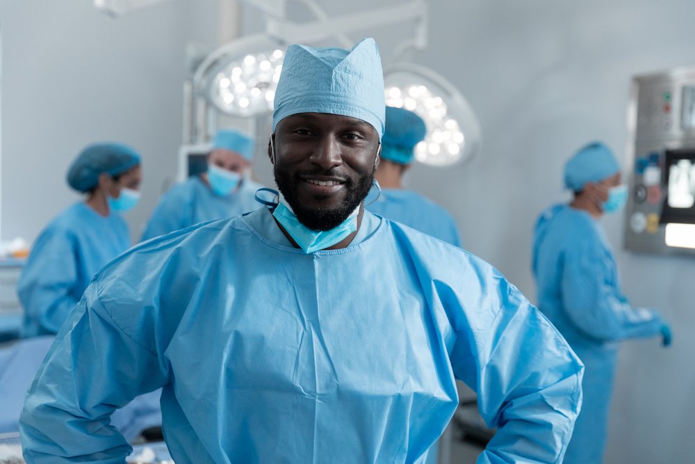 Smiling african american male surgeon with face mask and protective clothing in operating theatre. medicine, health and healthcare services during covid 19 coronavirus pandemic.